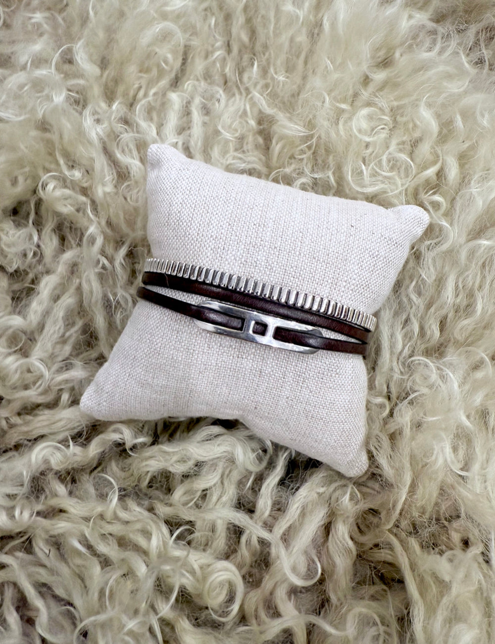 Bracelet stack featuring a ribbed sterling silver cuff and double-wrapped leather bracelet with sterling silver centerpiece on a neutral linen pillow.