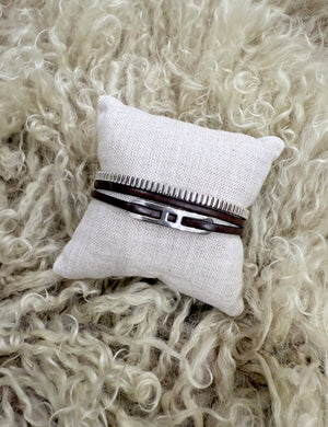 Bracelet stack featuring a ribbed sterling silver cuff and double-wrapped leather bracelet with sterling silver centerpiece on a neutral linen pillow.